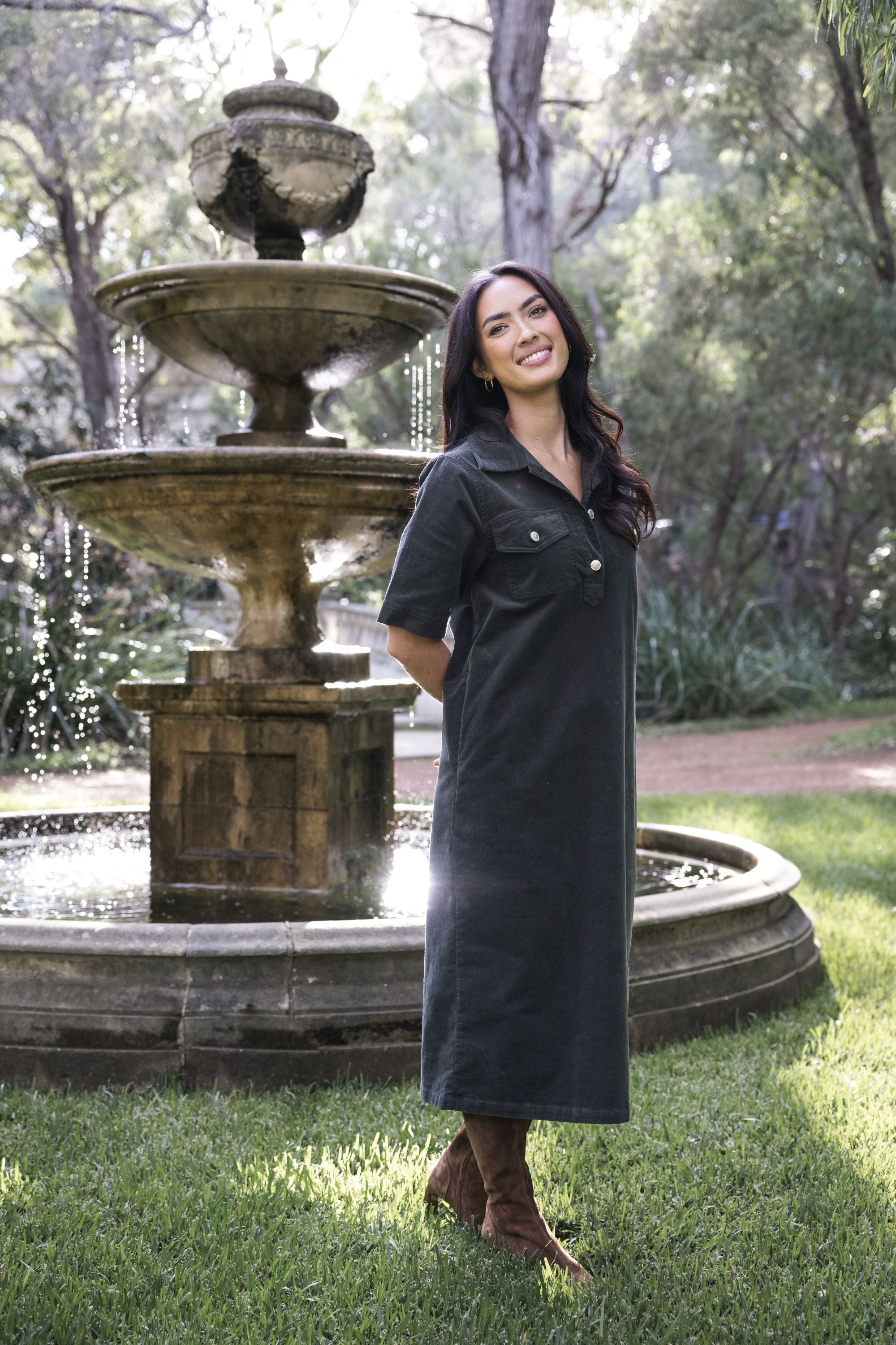 Model in lifestyle setting wearing Amici Cord Dress in Khaki with brown boots, full-length front view in front of a stone fountain in nature  