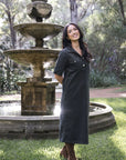 Model in lifestyle setting wearing Amici Cord Dress in Khaki with brown boots, full-length front view in front of a stone fountain in nature  