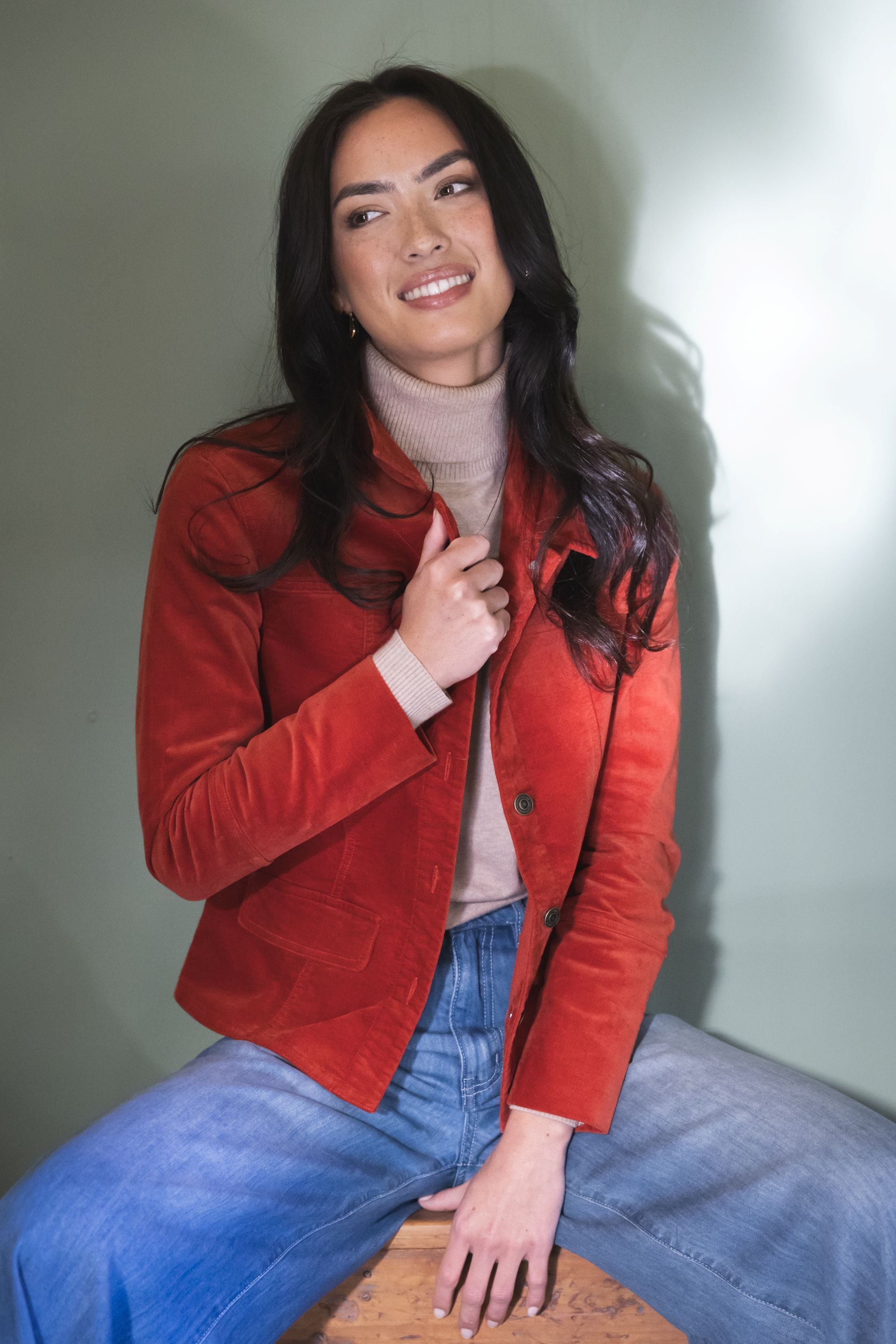 Model in lifestyle setting wearing Gabi Jacket in Rust over Alana Skivvy in Birch with Ryder Jean, close-up front view sitting 