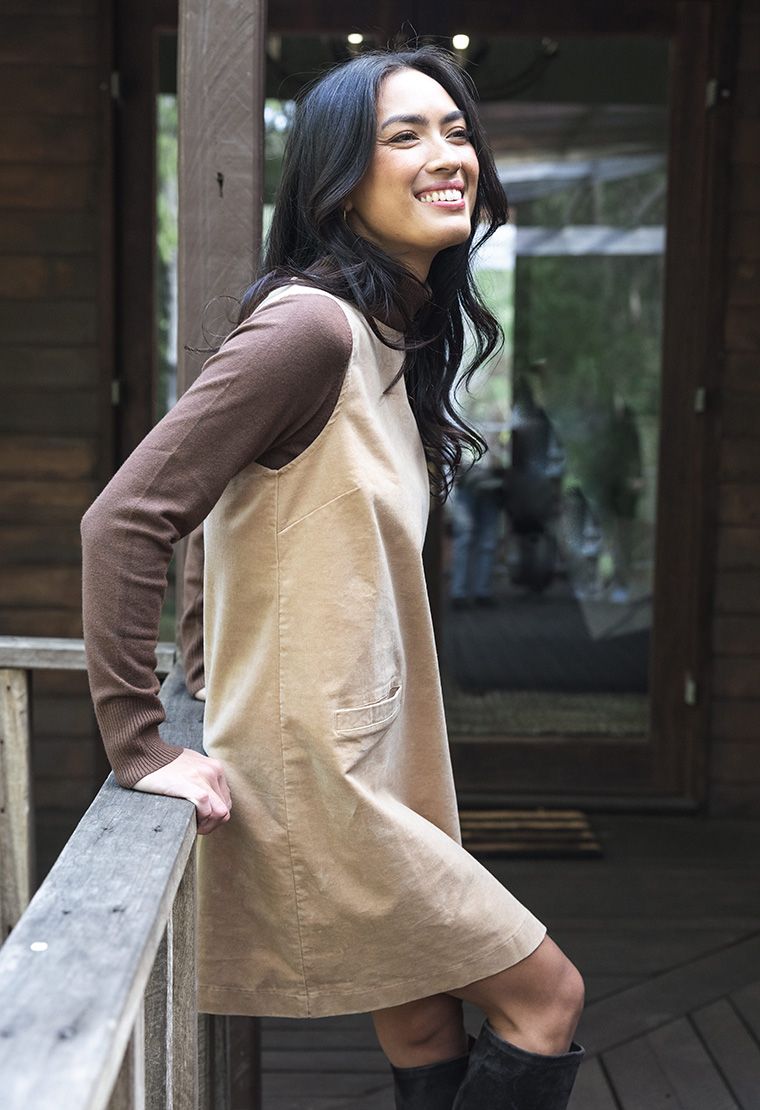 Model in lifestyle setting wearing Nova Cord Dress in Wheat over the Alana Skivvy in Chocolate, close-up side view leaning against wooden fence 