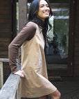 Model in lifestyle setting wearing Nova Cord Dress in Wheat over the Alana Skivvy in Chocolate, close-up side view leaning against wooden fence 
