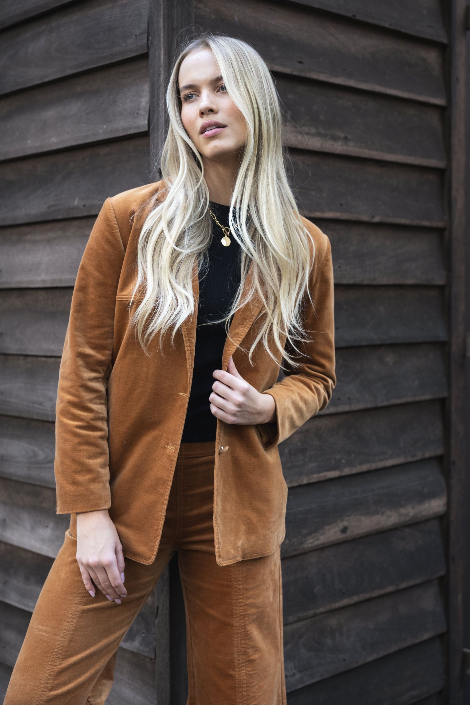 Model in lifestyle setting wearing Va Va Velour Coat in Camel over black top with matching Stitched Luxe Pant, close-up front view  