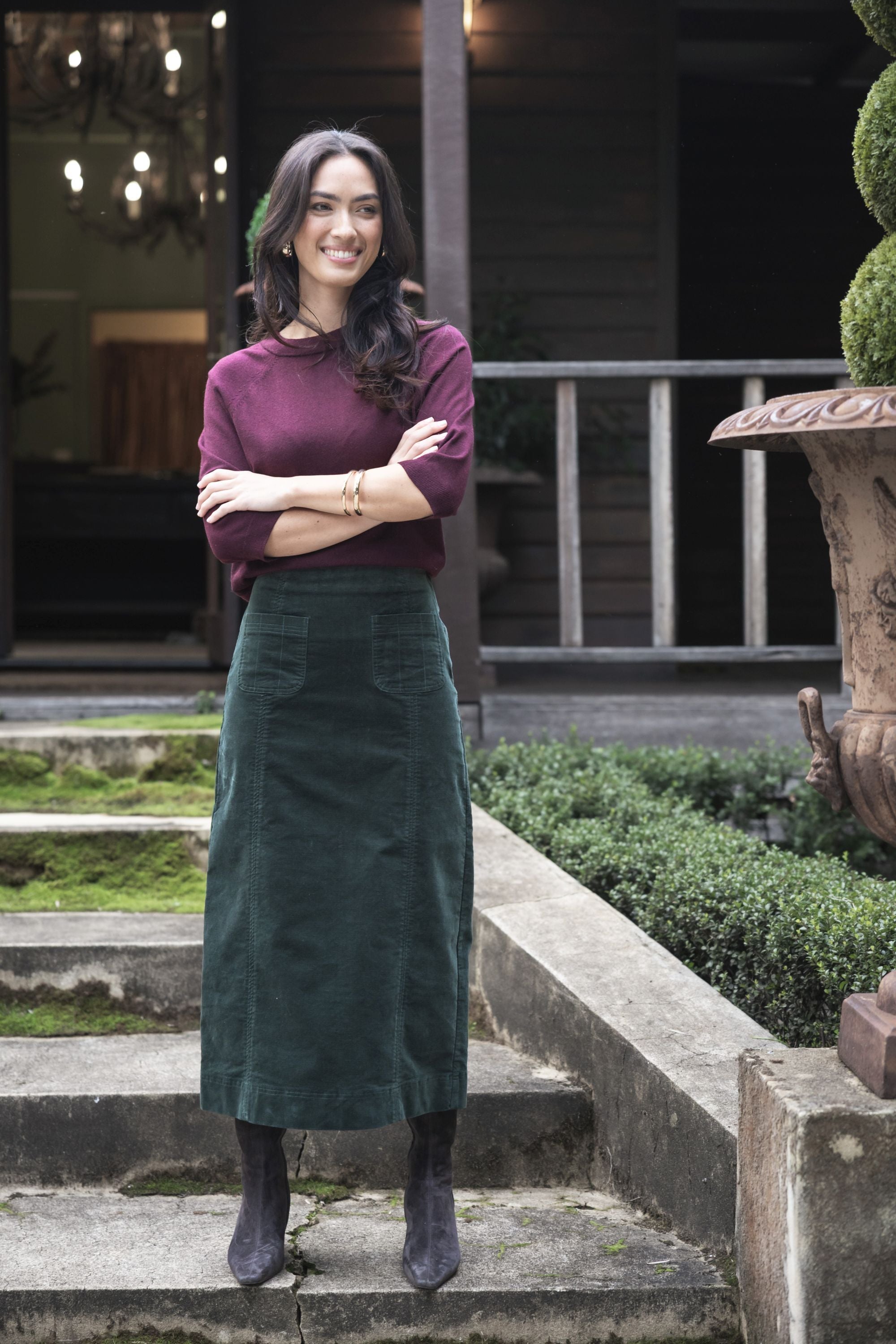 Model in lifestyle setting wearing Grace Top in Merlot with Va Va Velour Skirt in Pine, full-length front view on mossy stairs  