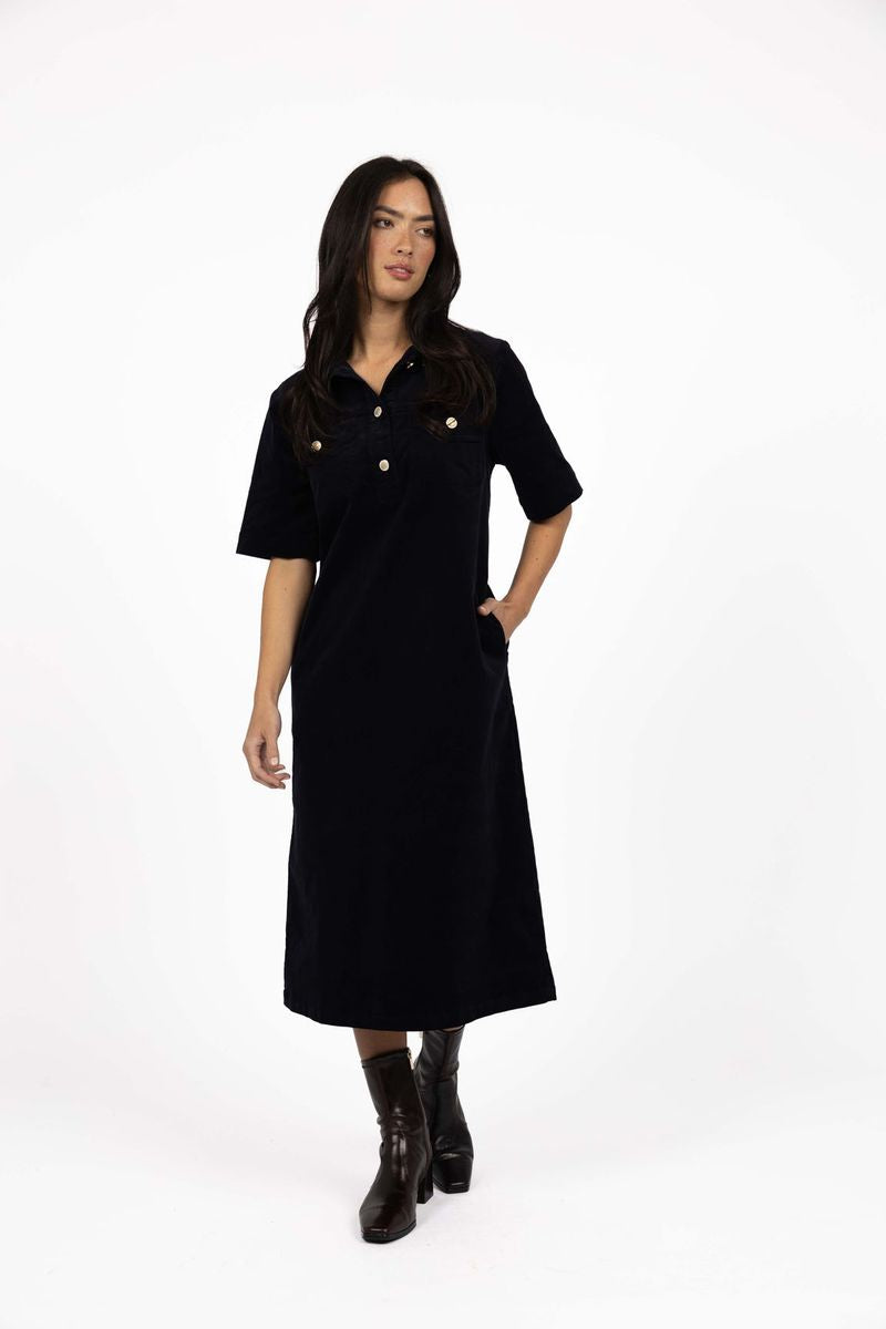 Model in studio wearing Amici Cord Dress in Indigo with brown boots, full-length front view with one hand in pocket  