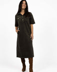 Model in studio wearing Amici Cord Dress in Khaki with brown boots, full-length front view with one hand in pocket  