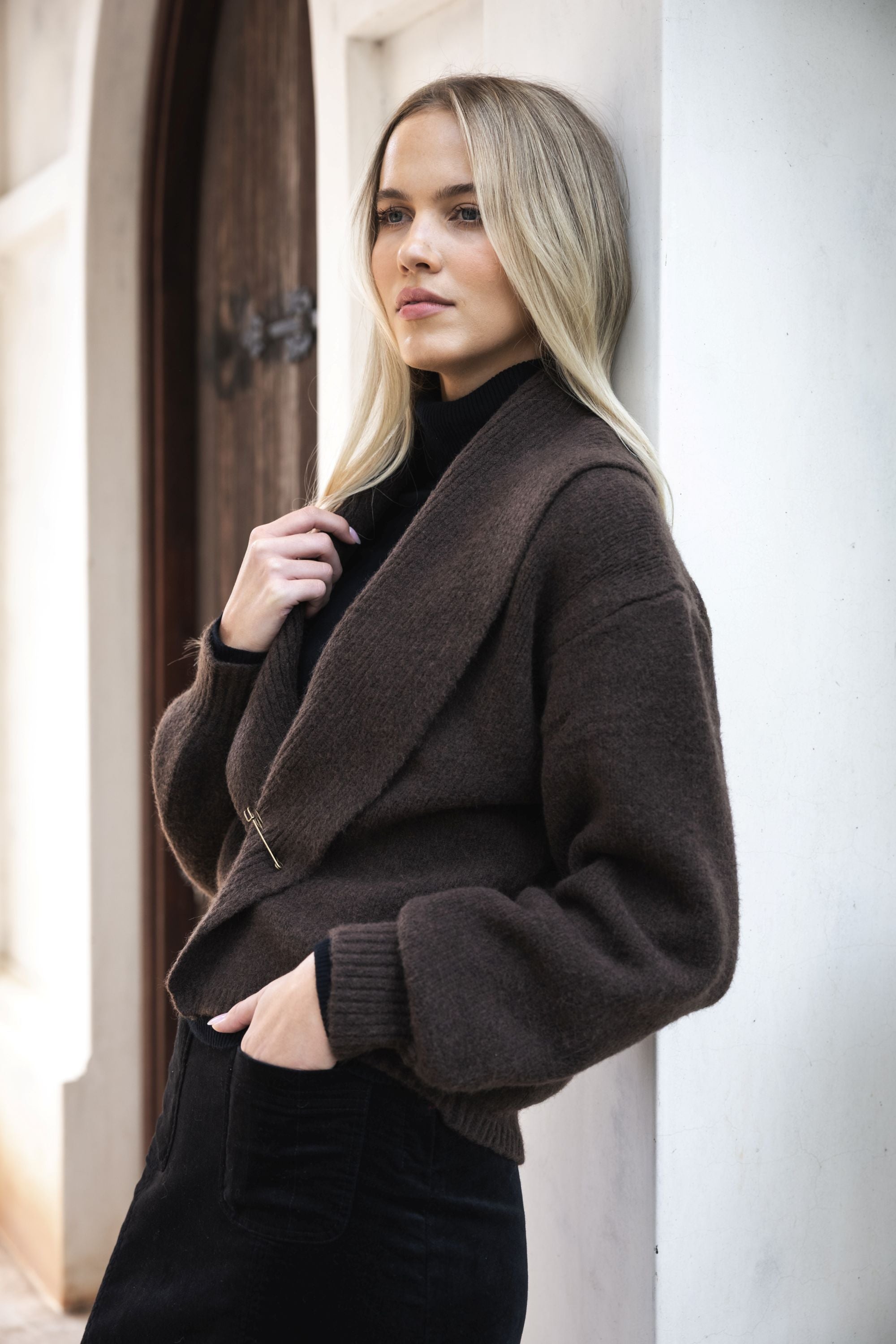 Model in lifestyle setting wearing knit Liana Cardi in Chocolate over black skivvy, close-up view leaning against a wall 