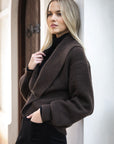 Model in lifestyle setting wearing knit Liana Cardi in Chocolate over black skivvy, close-up view leaning against a wall 