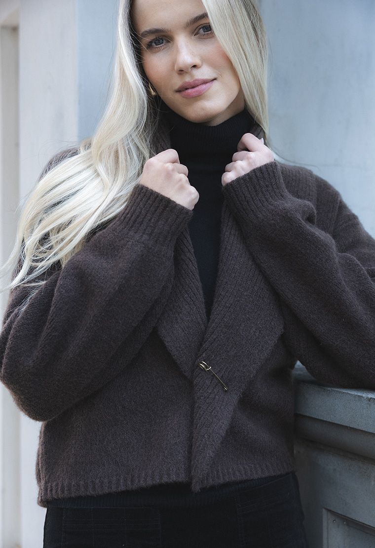 Model in lifestyle setting wearing Liana Cardi in Chocolate over black skivvy, close-up front view holding collar and showing brass pin  