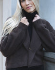 Model in lifestyle setting wearing Liana Cardi in Chocolate over black skivvy, close-up front view holding collar and showing brass pin  