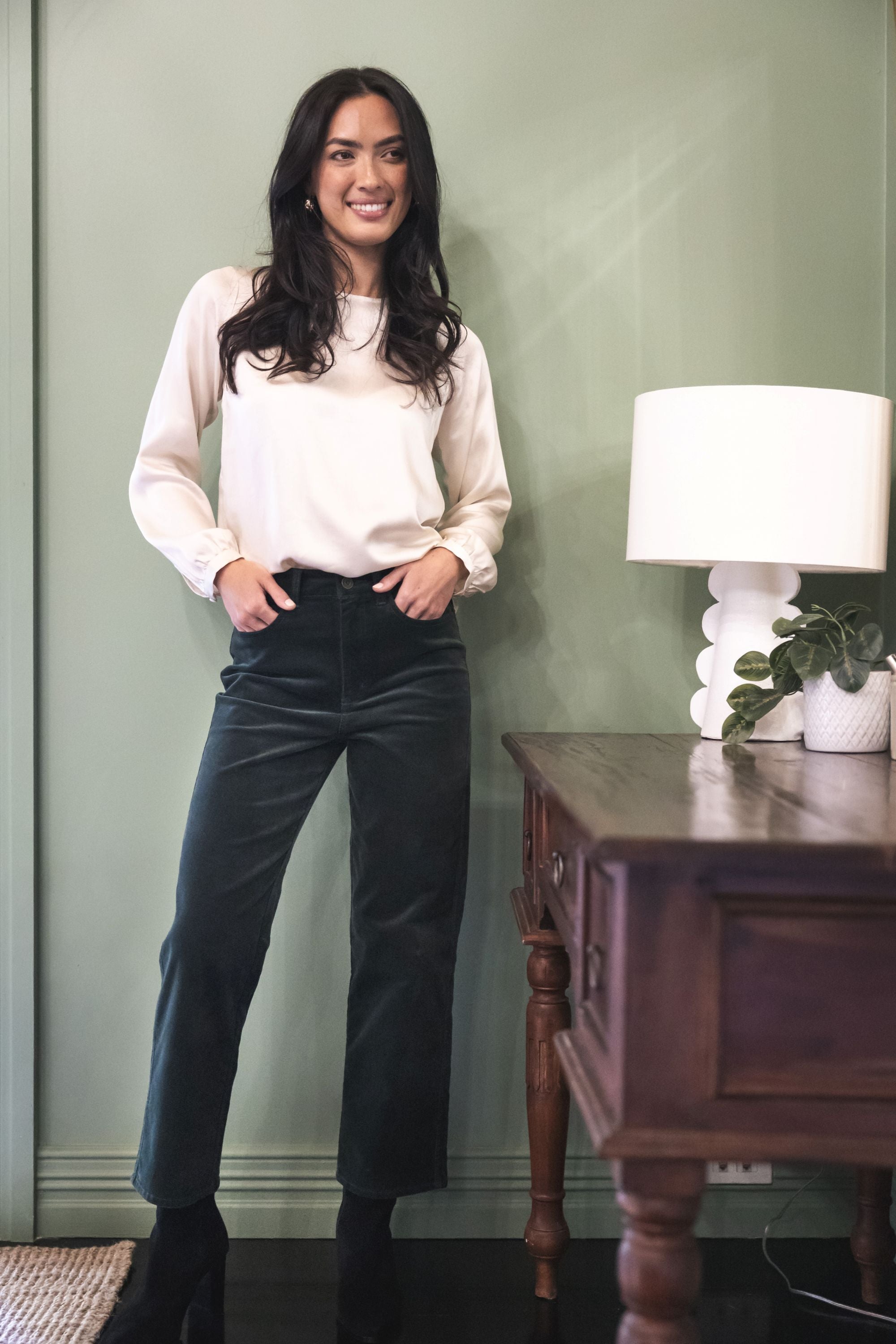 Model in lifestyle setting wearing Va Va Velour Pant in Pine with Alina Top in Cream, full-length front view  