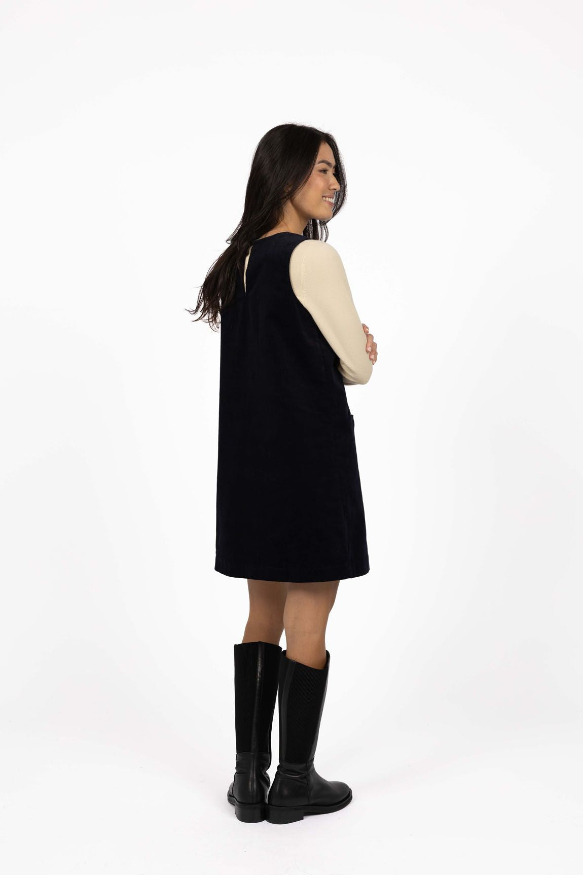Model in studio wearing Nova Cord Dress in Indigo over the Mae Jumper in Buttermilk with black boots, full-length back view 